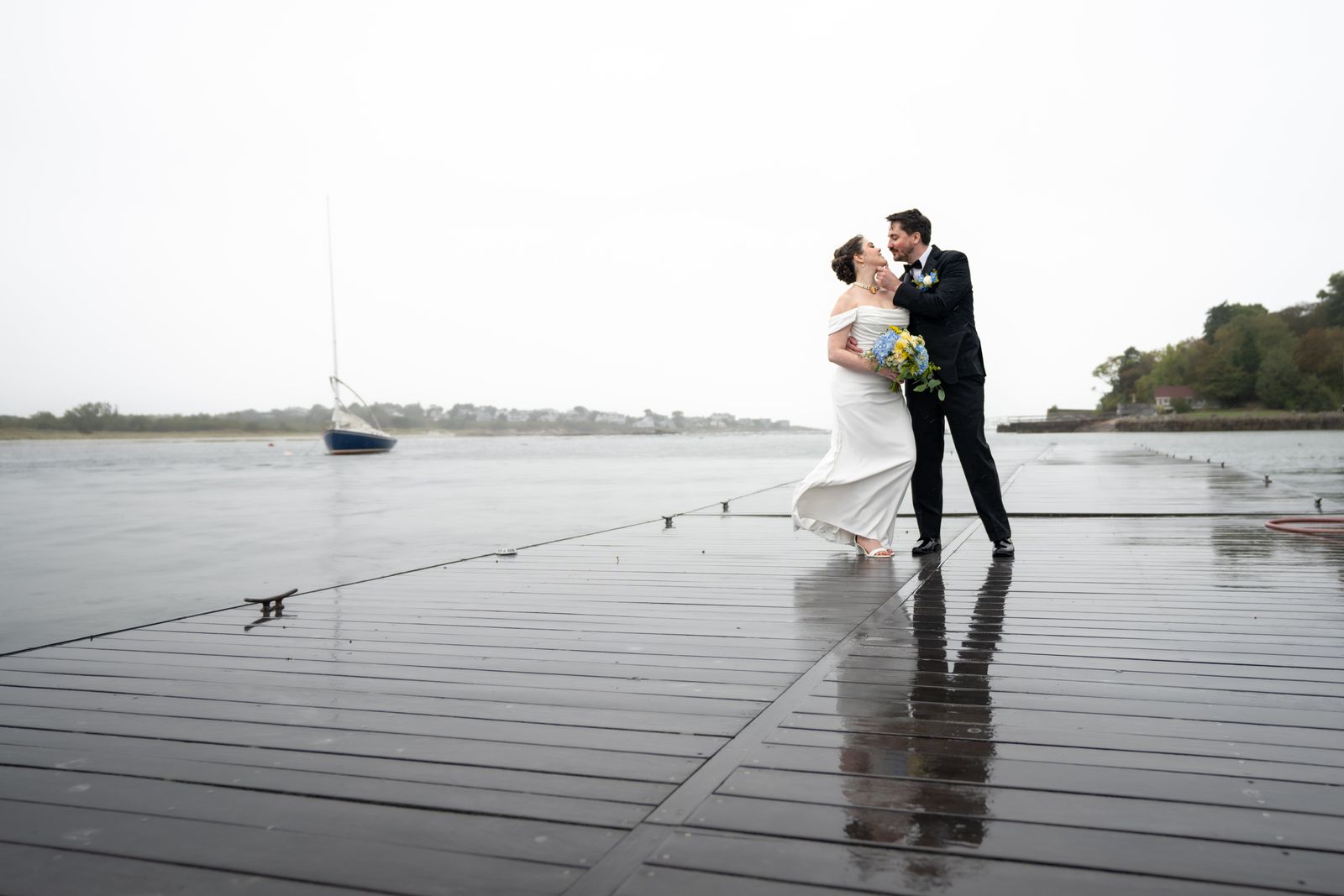 Couple kissing on a long rain-slicked dock with a sailboat moored in the harbor